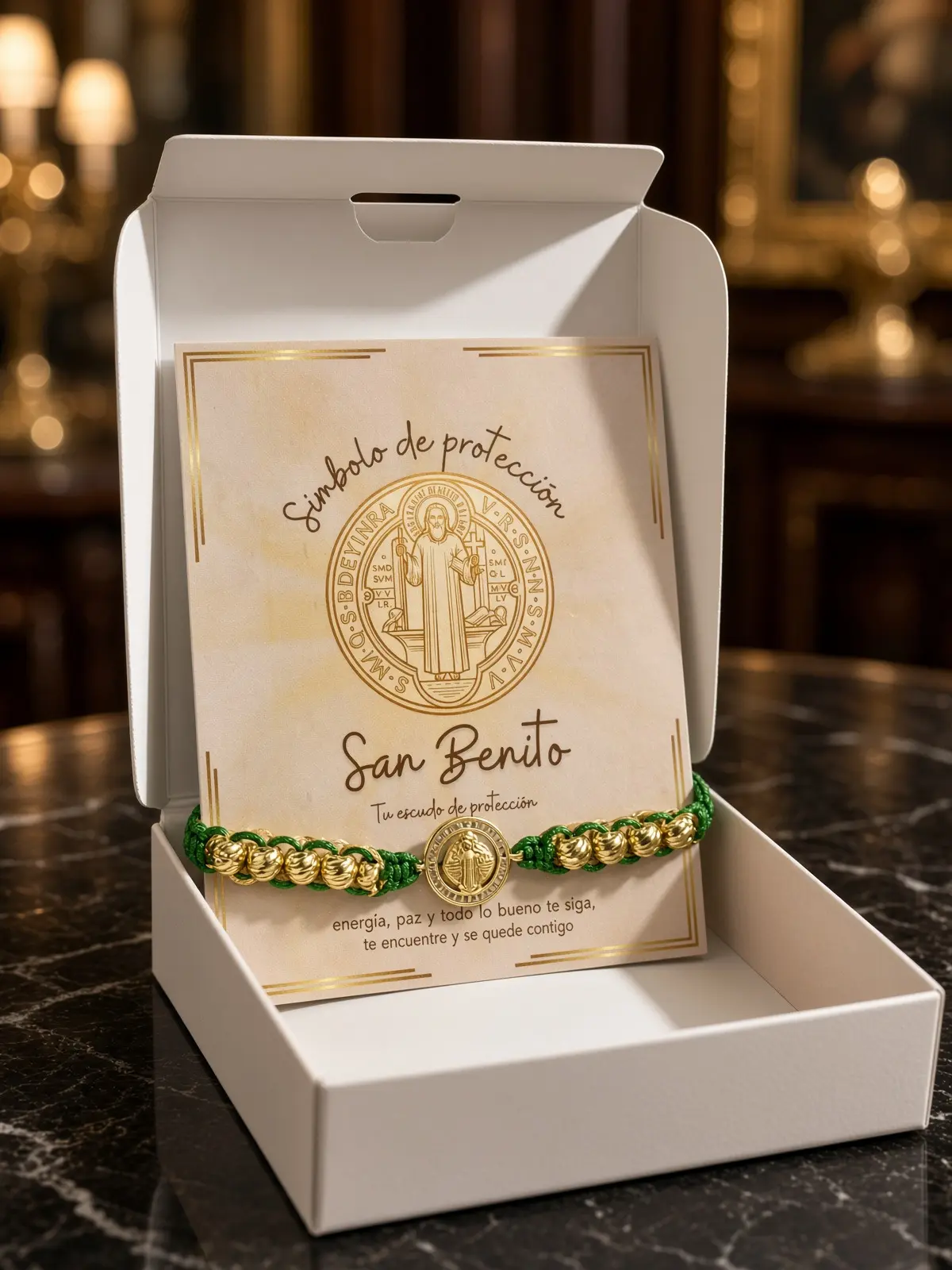 Pulsera tejida verde con cuentas y medalla dorada de San Benito dentro de caja blanca con tarjeta de protección.