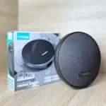 Parlante portátil Bluetooth Treqa gris oscuro redondo con tela texturizada y su caja de empaque sobre superficie de madera.