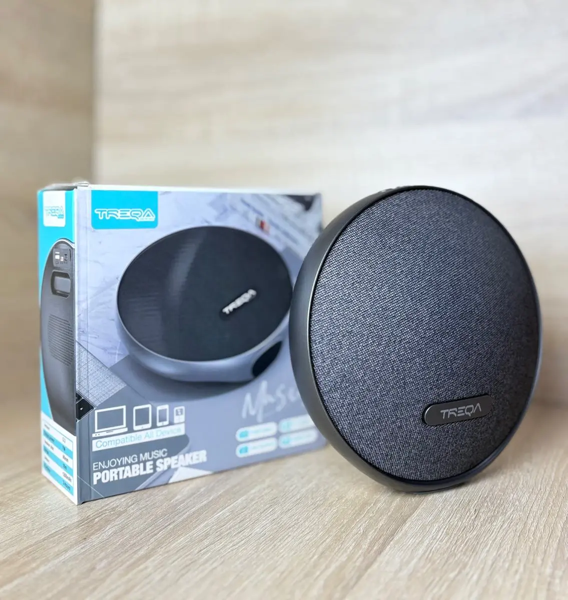 Parlante portátil Bluetooth Treqa gris oscuro redondo con tela texturizada y su caja de empaque sobre superficie de madera.