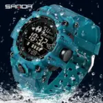 Reloj táctico deportivo digital Sanda en color verde azulado sumergido parcialmente en agua, diseño robusto de alta resistencia y pantalla digital iluminada.