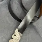 Correa de cuero negro con interior café y hebilla metálica con diseño de alas, mostrada en ángulo sobre una superficie gris texturizada.