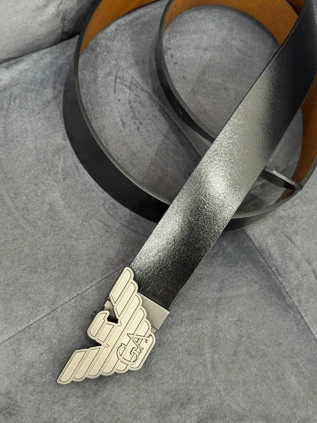 Correa de cuero negro con interior café y hebilla metálica con diseño de alas, mostrada en ángulo sobre una superficie gris texturizada.