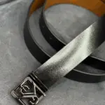 Correa de cuero negro con textura granulada, interior color canela y hebilla metálica con diseño geométrico entrelazado.