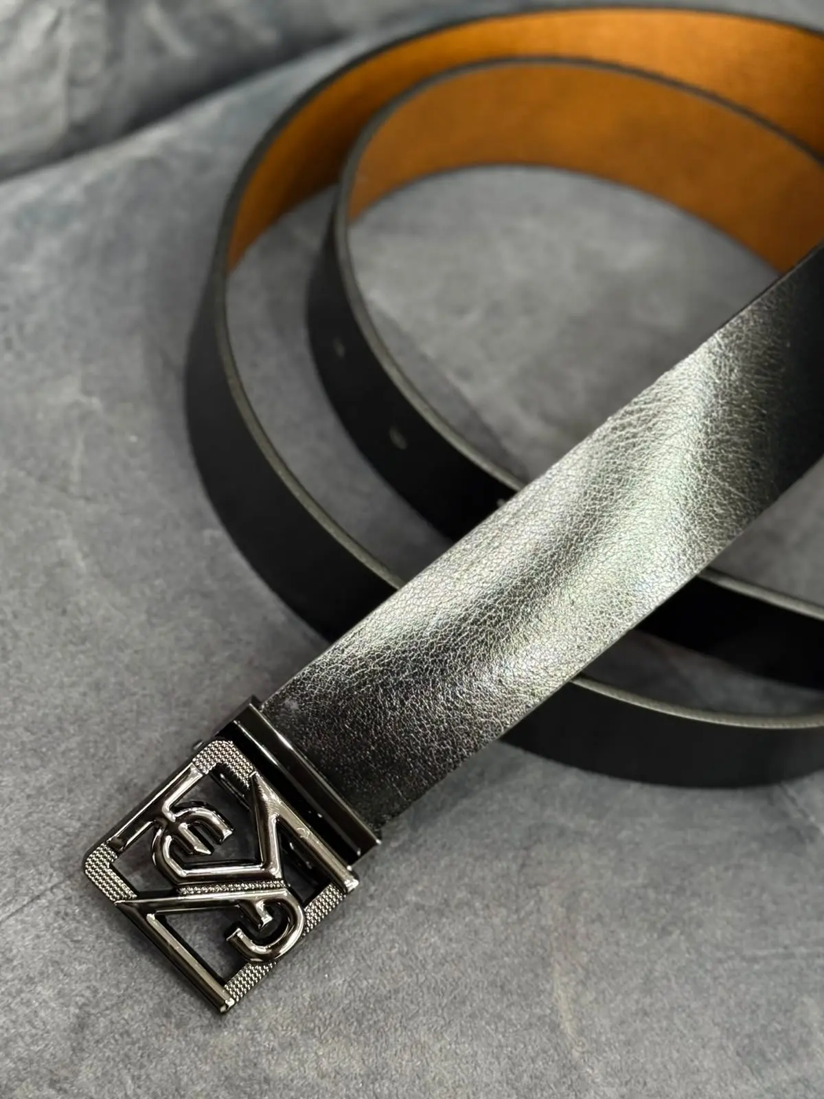 Correa de cuero negro con textura granulada, interior color canela y hebilla metálica con diseño geométrico entrelazado.