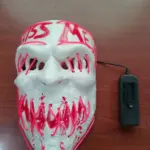 Máscara blanca estilo The Purge con luces LED rosadas, texto KISS ME y diseño de dientes, incluye caja de baterías negra sobre fondo de madera.