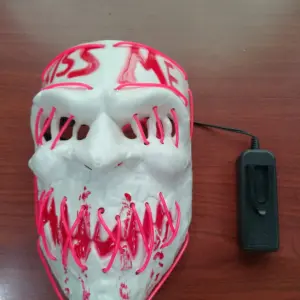 Máscara blanca estilo The Purge con luces LED rosadas, texto KISS ME y diseño de dientes, incluye caja de baterías negra sobre fondo de madera.