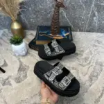 Sandalias negras de plataforma con doble correa decorada con pedrería brillante y hebillas plateadas, sostenidas en una mano.
