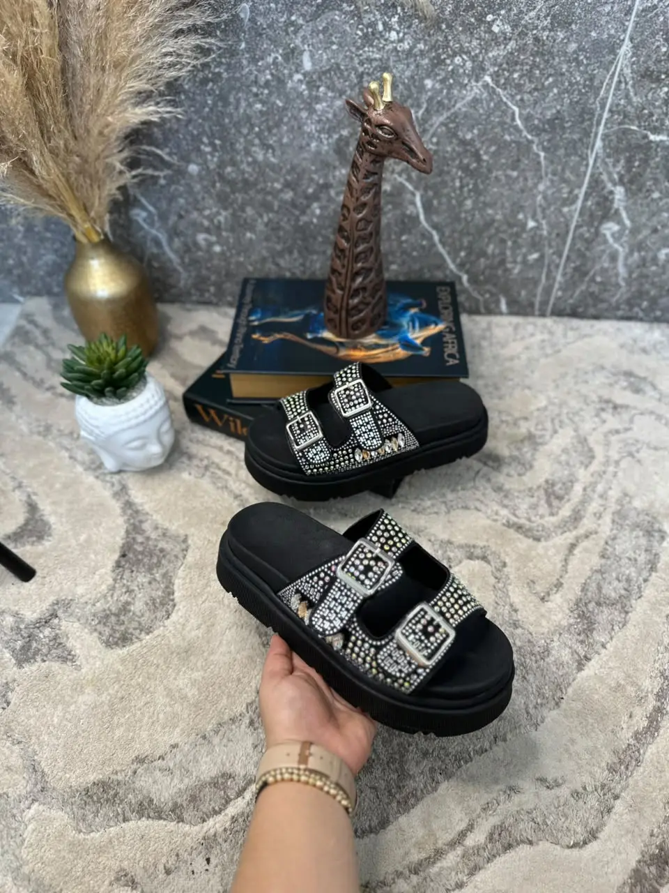 Sandalias negras de plataforma con doble correa decorada con pedrería brillante y hebillas plateadas, sostenidas en una mano.