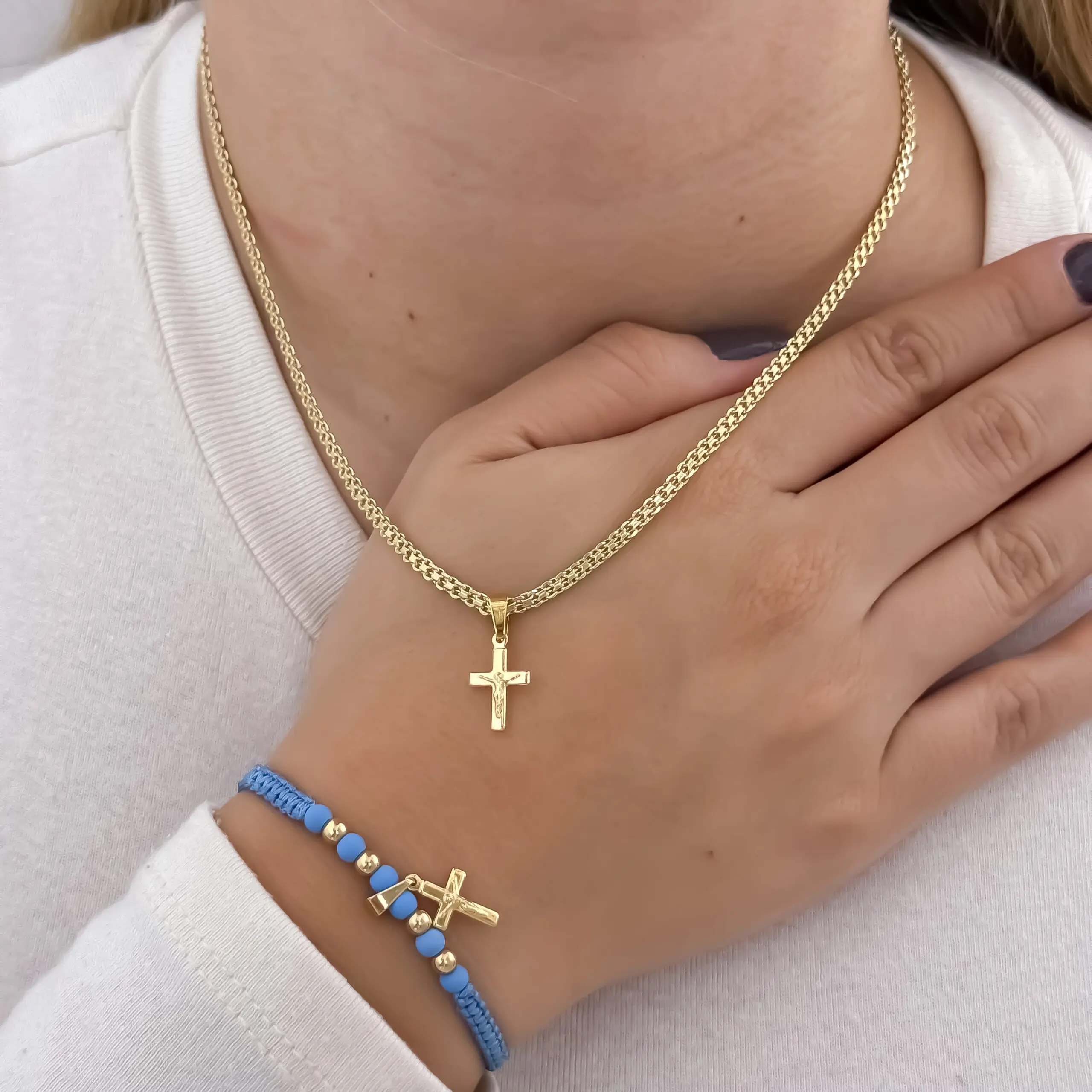 Conjunto de collar tejido y manilla azul con abalorios dorados, ambos con dije de cruz en oro laminado de 18K, puestos en el pecho.