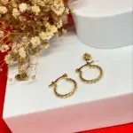 Par de candongas de oro laminado con diseño trenzado tipo melcocha de 1.5 cm, sobre un pedestal blanco con flores secas.