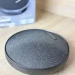 Parlante Bluetooth portátil circular color gris oscuro con cubierta de tela texturizada sobre superficie de madera y su caja de empaque al fondo.
