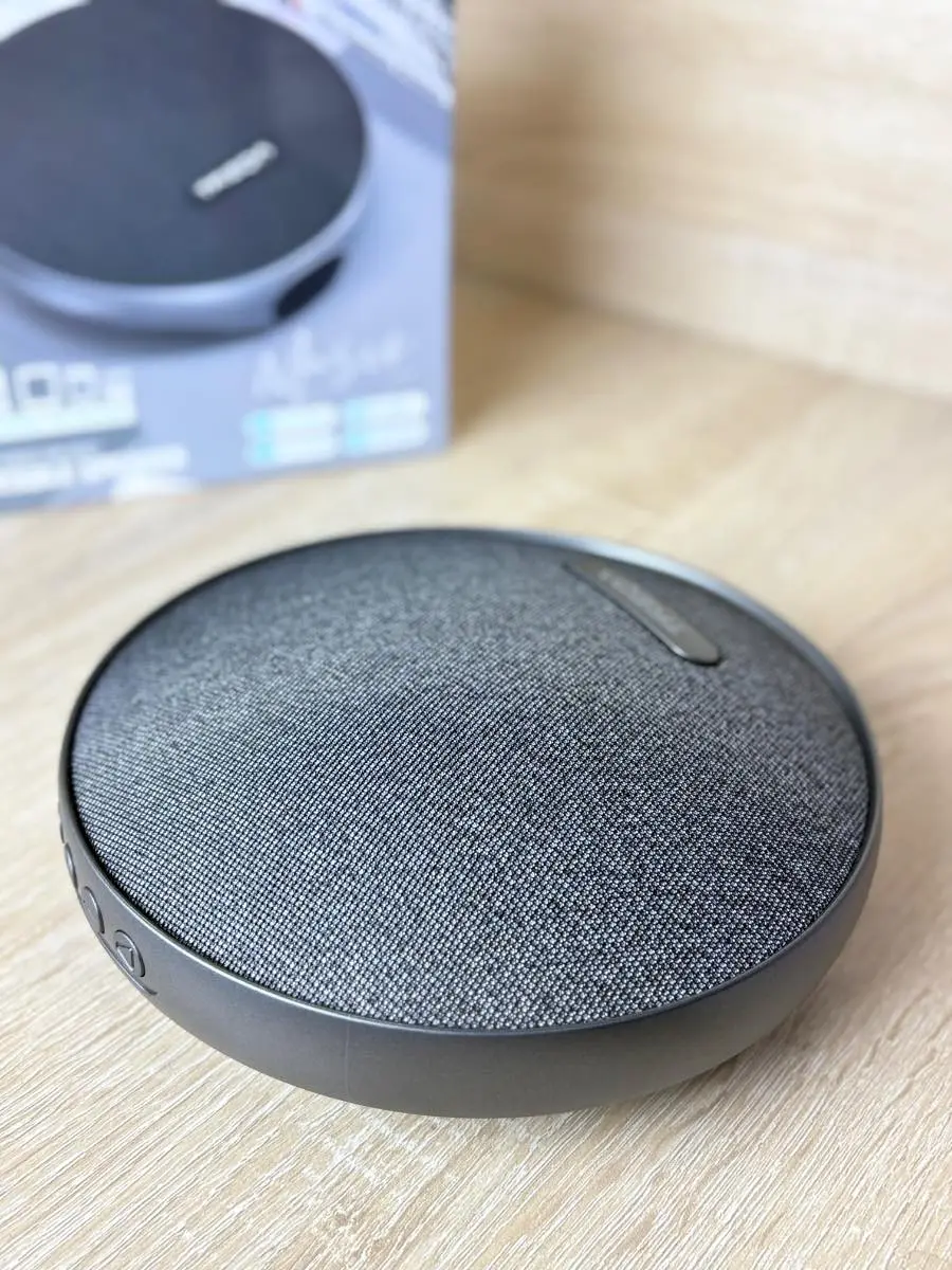 Parlante Bluetooth portátil circular color gris oscuro con cubierta de tela texturizada sobre superficie de madera y su caja de empaque al fondo.