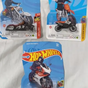 Tres motos de juguete Hot Wheels en sus empaques de cartón azul; una cruiser marrón, una de motocross naranja y una deportiva roja.