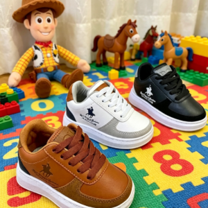 Zapatos tipo polo casual para niño en color miel con suela blanca, acompañados de otras dos variantes en blanco y negro, sobre alfombra didáctica junto a juguetes.
