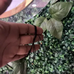 Set de dos pulseras ajustables de cordón negro con pequeños detalles metálicos dorados, sostenidas por una mano sobre un fondo de follaje verde.