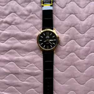 Reloj Orient de cuarzo con esfera negra, caja y marcadores dorados, doble calendario y pulsera de cuero negro con textura de cocodrilo sobre fondo acolchado rosado.