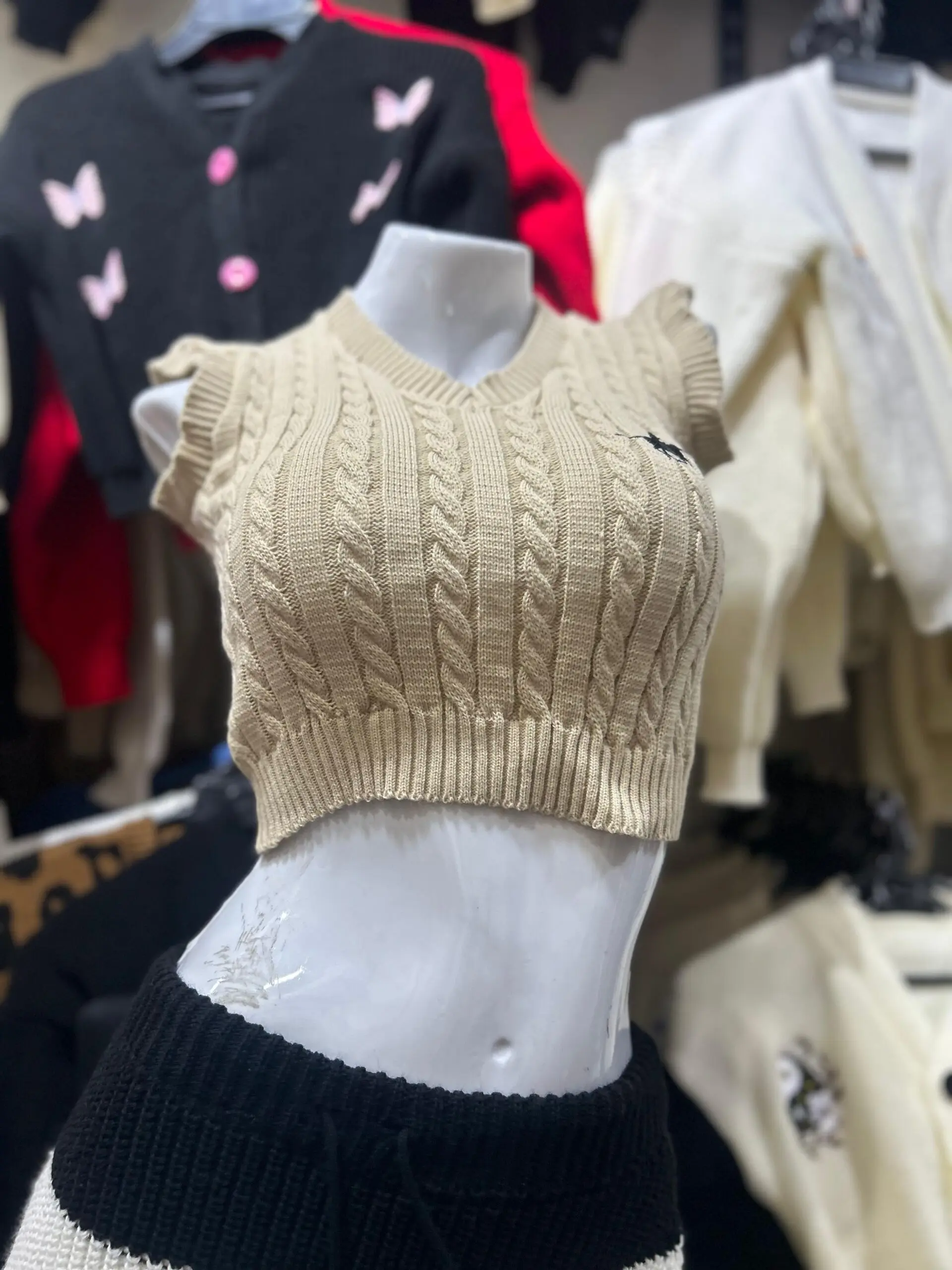 Top tejido sin mangas color beige con cuello en V, diseño de trenzas y volantes en los hombros, mostrado en un maniquí blanco.