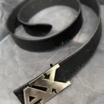 Correa de cuero negra para hombre con hebilla metálica grande con las letras AX, mostrada en ángulo sobre fondo gris texturizado.
