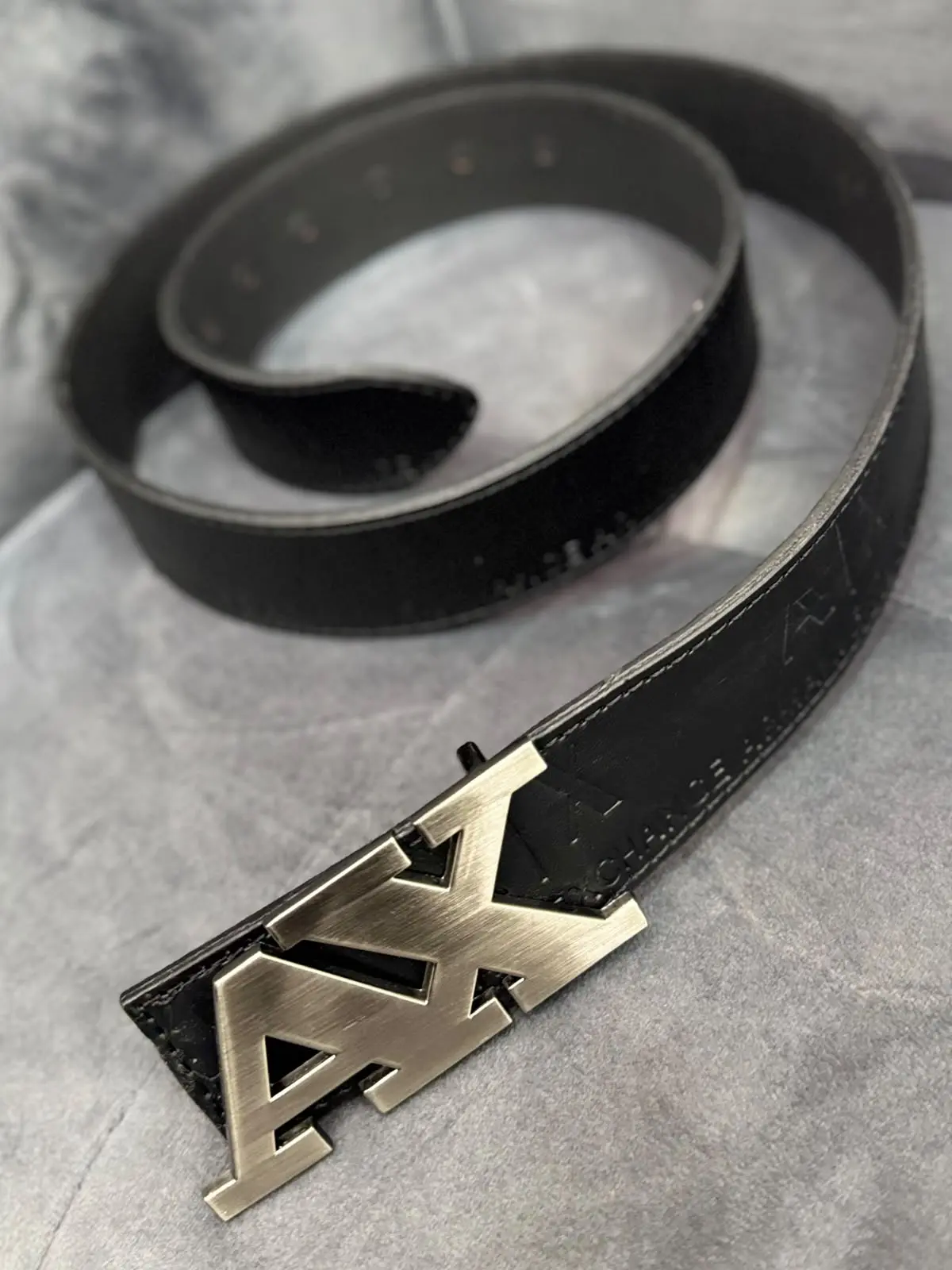 Correa de cuero negra para hombre con hebilla metálica grande con las letras AX, mostrada en ángulo sobre fondo gris texturizado.