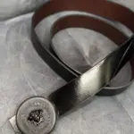 Correa de cuero negra para hombre con forro marrón y hebilla metálica circular con diseño en relieve sobre fondo gris.