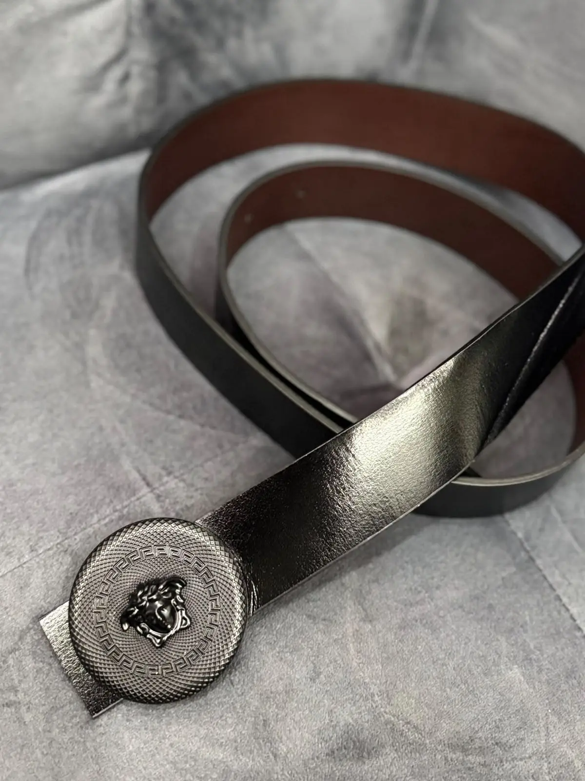 Correa de cuero negra para hombre con forro marrón y hebilla metálica circular con diseño en relieve sobre fondo gris.