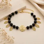 Pulsera de piedra ónix negra con abalorios dorados en forma de hojas y dije central de rosa en acero inoxidable sobre fondo neutro.