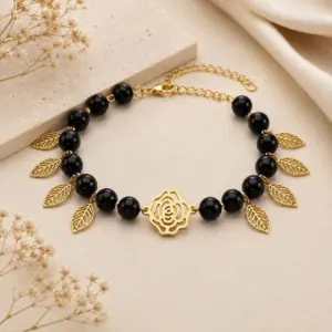 Pulsera de piedra ónix negra con abalorios dorados en forma de hojas y dije central de rosa en acero inoxidable sobre fondo neutro.