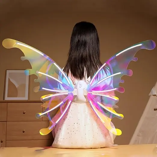 Niña de espaldas con alas de hada iridiscentes, luces LED blancas y rosadas, y motor central blanco para disfraz infantil en habitación.