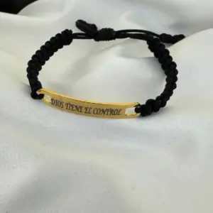 Pulsera ajustable de cordón negro trenzado con placa de oro de 18k grabada con la frase Dios Tiene El Control sobre fondo de tela blanca.
