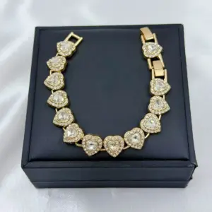Pulsera dorada con dijes de corazones cristalinos pavé dispuesta sobre estuche negro cuadrado para regalo elegante.