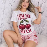Mujer rubia sentada luciendo pijama blanca de manga corta con estampado de cerezas y short rosa a cuadros sobre base peluda.