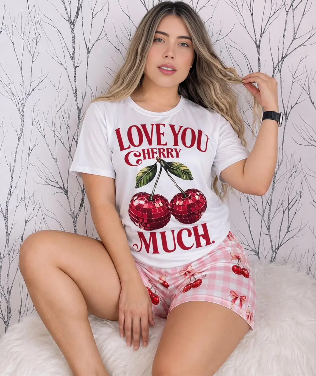 Mujer rubia sentada luciendo pijama blanca de manga corta con estampado de cerezas y short rosa a cuadros sobre base peluda.