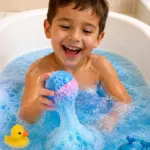 Niño jugando en bañera con agua azul y espuma, usando bomba de baño azul y rosa efervescente junto a juguetes de pato y ballena.