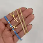 Mano sosteniendo conjunto dorado de dos piezas: collar tejido con dije de cruz y manilla azul ajustable con abalorios y cruz en oro laminado de 18K.