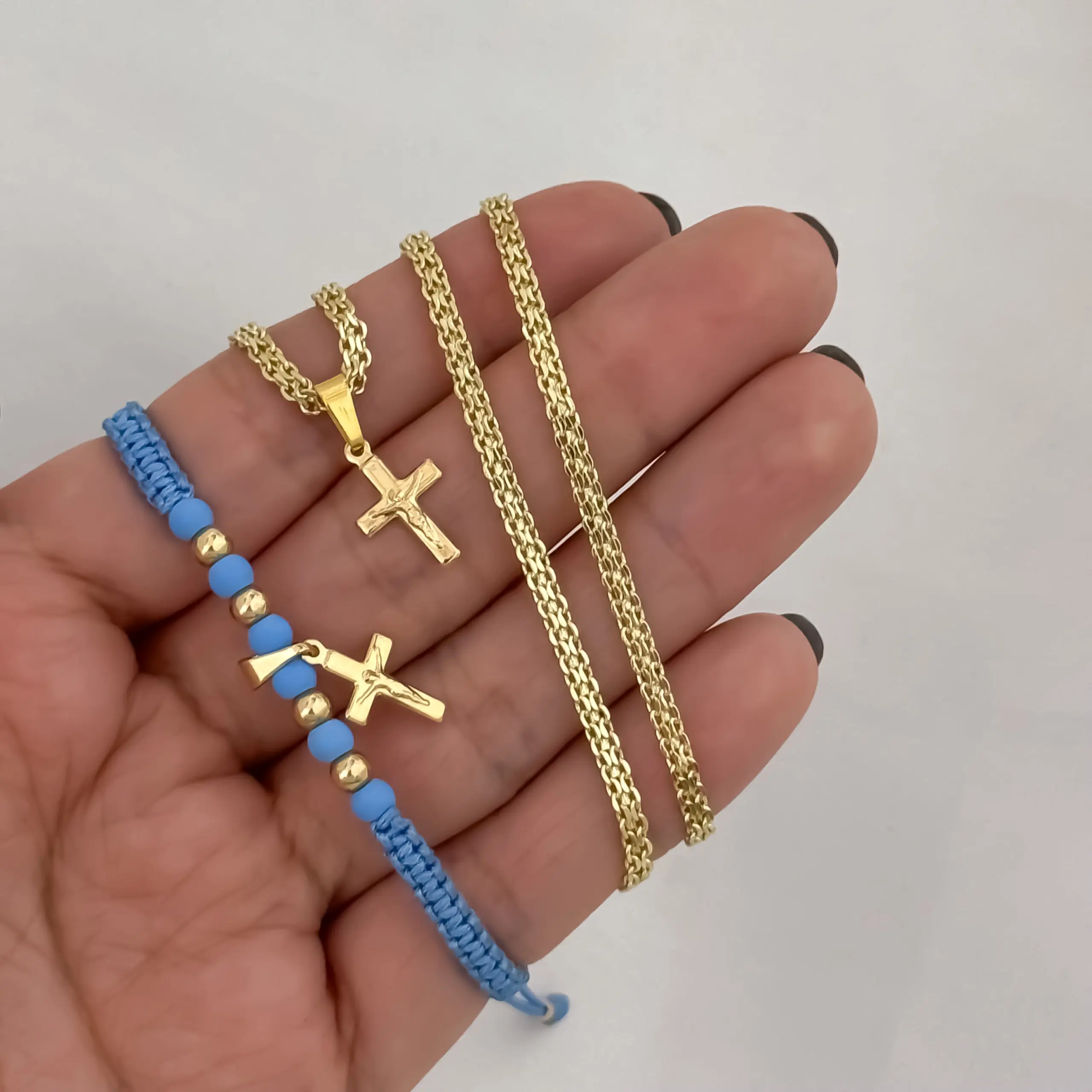 Mano sosteniendo conjunto dorado de dos piezas: collar tejido con dije de cruz y manilla azul ajustable con abalorios y cruz en oro laminado de 18K.