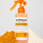 Spray exfoliante corporal naranja con ácido kójico y cúrcuma, flanqueado por montículos de polvos amarillo y blanco sobre superficie blanca.