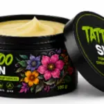 Tarro negro de 100 g de pomada Tattoo Skin con crema de textura suave color crema, envase con diseño floral y tapa apoyada al lado.