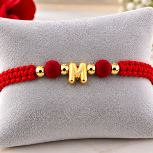 Pulsera de hilo rojo trenzado con inicial "M" central en oro laminado de 18K, adornada con cuentas doradas y esferas aterciopeladas sobre cojín gris.