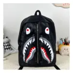 Mochila escolar negra impermeable con diseño de cara de tiburón, ojos rojos y boca abierta con dientes blancos, vista frontal.
