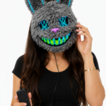 Máscara de conejo gris de peluche con luces LED azules en orejas, ojos y boca verde fluorescente, sostenida por mujer con control.