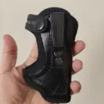 Funda negra para pistola en cuero ecológico con gancho de sujeción metálico, costuras reforzadas y correa con botón a presión sobre mano humana