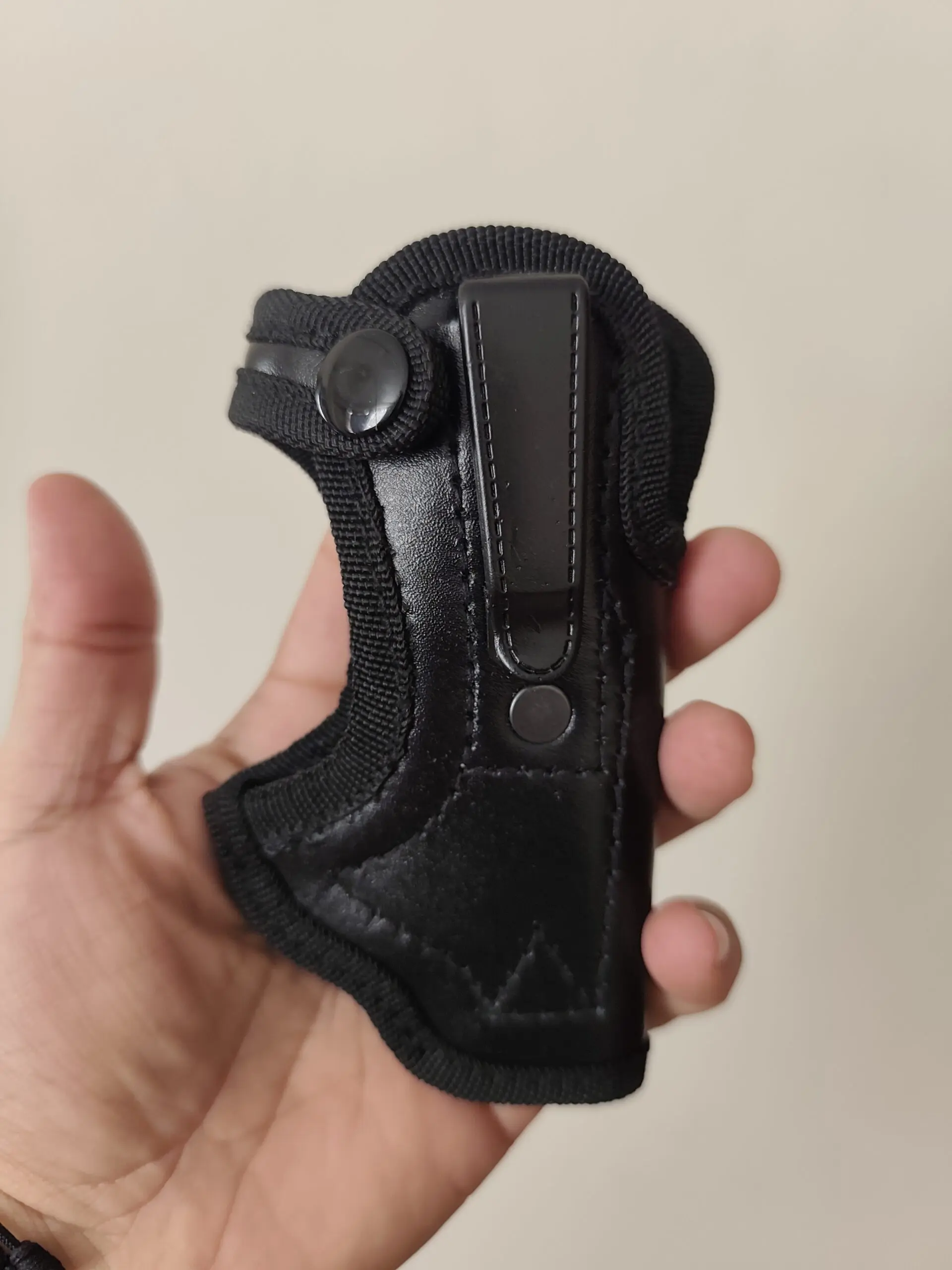 Funda negra para pistola en cuero ecológico con gancho de sujeción metálico, costuras reforzadas y correa con botón a presión sobre mano humana