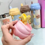 Mano sosteniendo la tapa de un termo kawaii rosa con clip amarillo, frente a varios termos coloridos de diseño infantil.