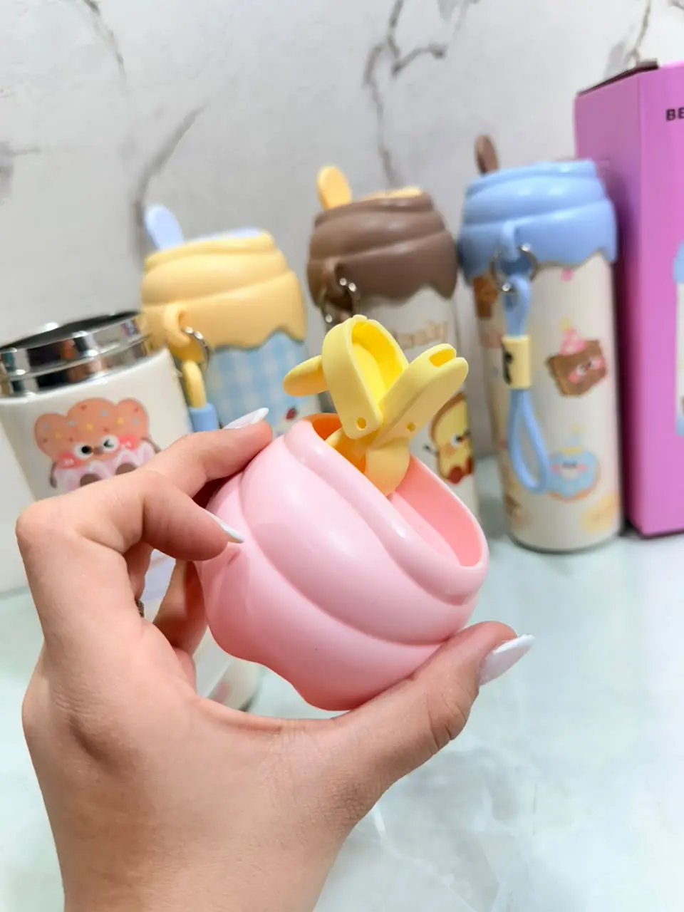 Mano sosteniendo la tapa de un termo kawaii rosa con clip amarillo, frente a varios termos coloridos de diseño infantil.