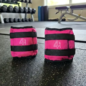 Par de pesas de 4 kg color rosa con correas de velcro negras para tobillos o muñecas, sobre piso de gimnasio con mancuernas al fondo.