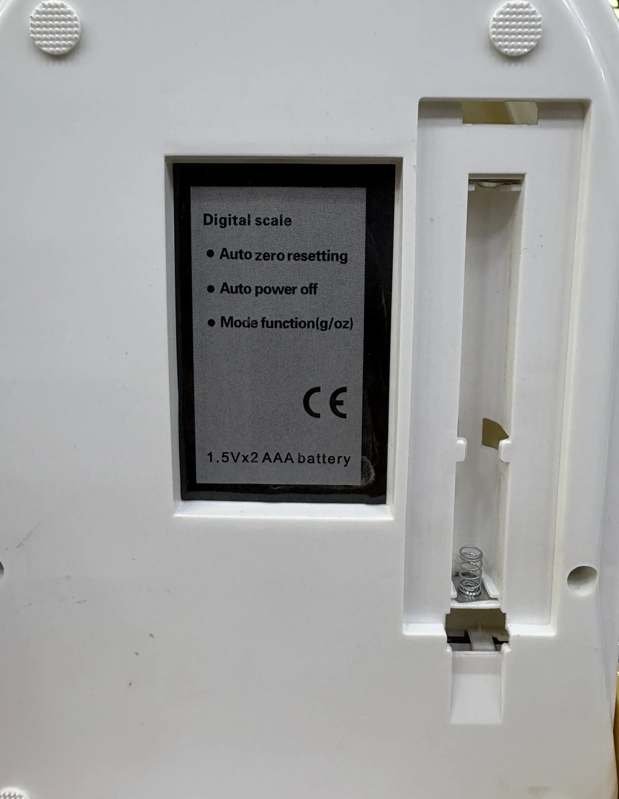 Parte inferior de báscula gramera digital blanca en plástico con compartimiento abierto para dos baterías AAA y etiqueta técnica gris.