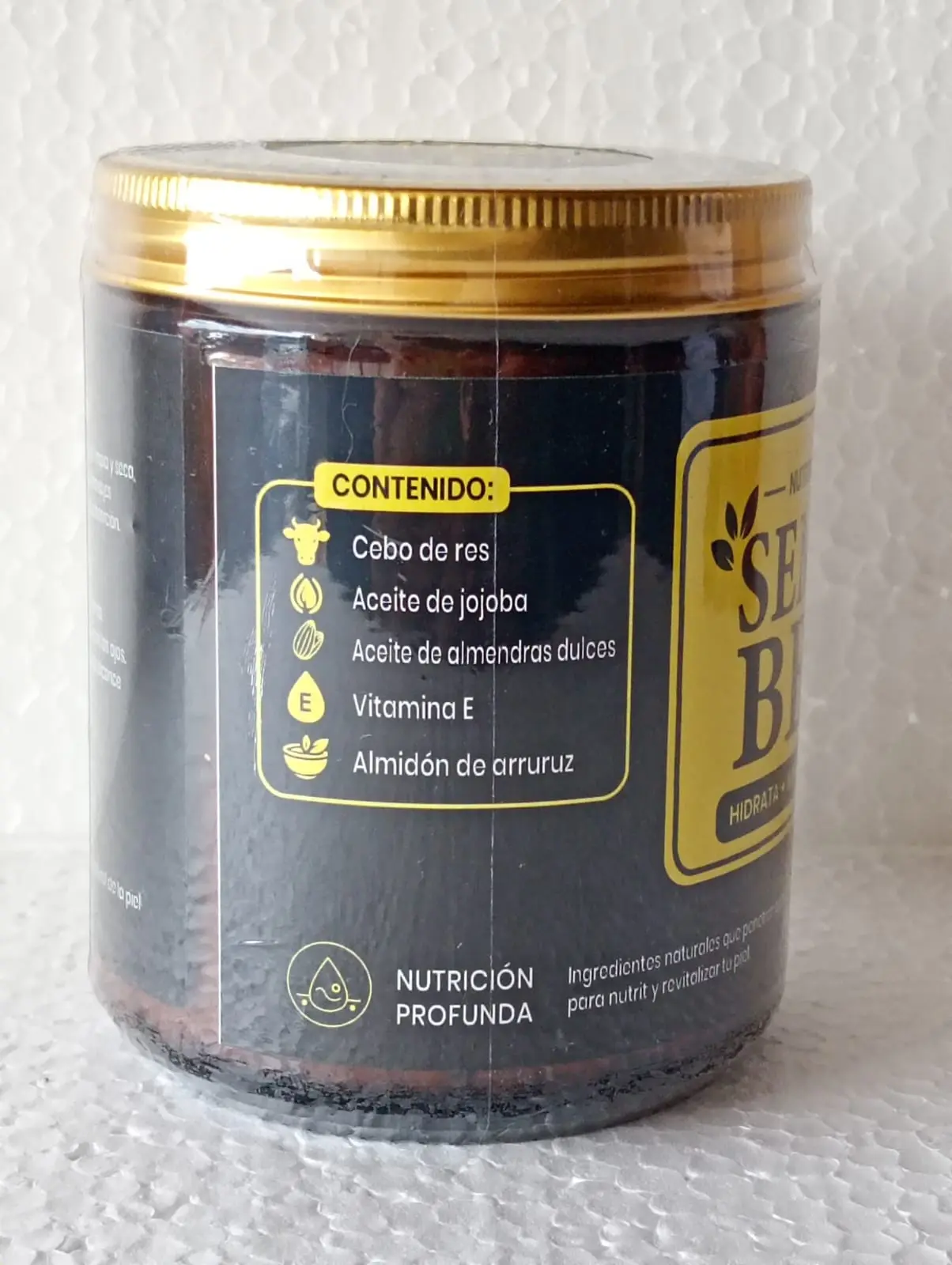 Tarro de vidrio ámbar con tapa dorada que muestra la lista de ingredientes naturales en su etiqueta negra con bordes amarillos.