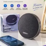 Parlante Bluetooth portátil circular gris con acabado en tela, junto a su caja, un teléfono inteligente reproduciendo música y el texto promocional superior.
