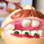 Modelo de dentadura didáctica infantil en plástico beige con dientes y lengua de masa moldeable que simula caries con manchas verdes.