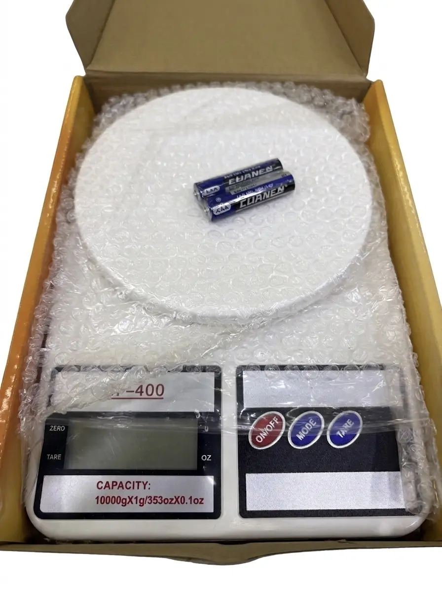 Báscula gramera digital blanca para cocina dentro de su caja de cartón, con dos pilas AA sobre la plataforma circular y pantalla digital con capacidad hasta de 10000 gramos